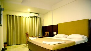Habitación de hotel con una cama grande, cabecera de madera, una silla, una mesa con una botella y una cortina.