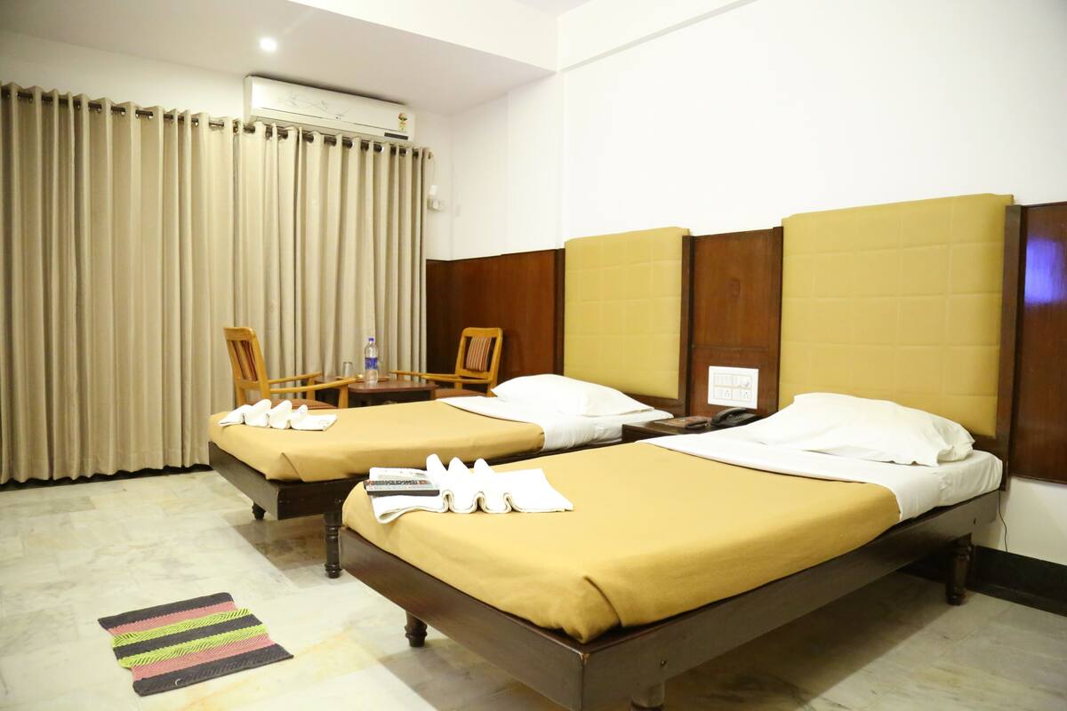 A hotel room with two beds, a wooden headboard, a small table with a chair, and a window with curtains.