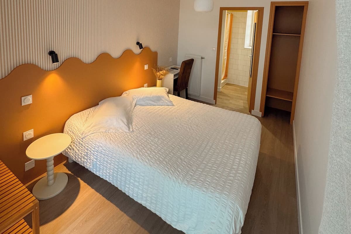 A hotel room with a bed, bedside table, lamp, and a view of the hallway.