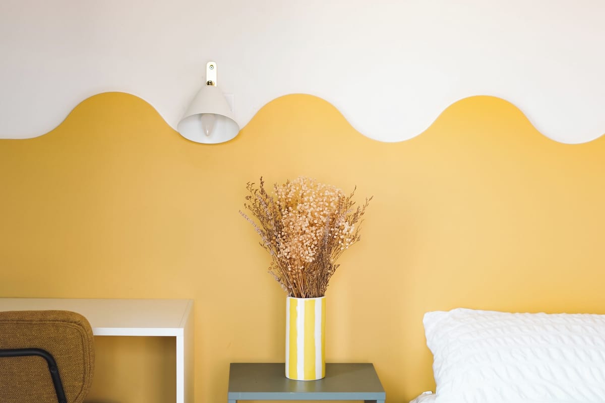 A room with a yellow accent wall, a white bed, a small table with a vase of dried flowers, a chair, and a desk with a lamp.