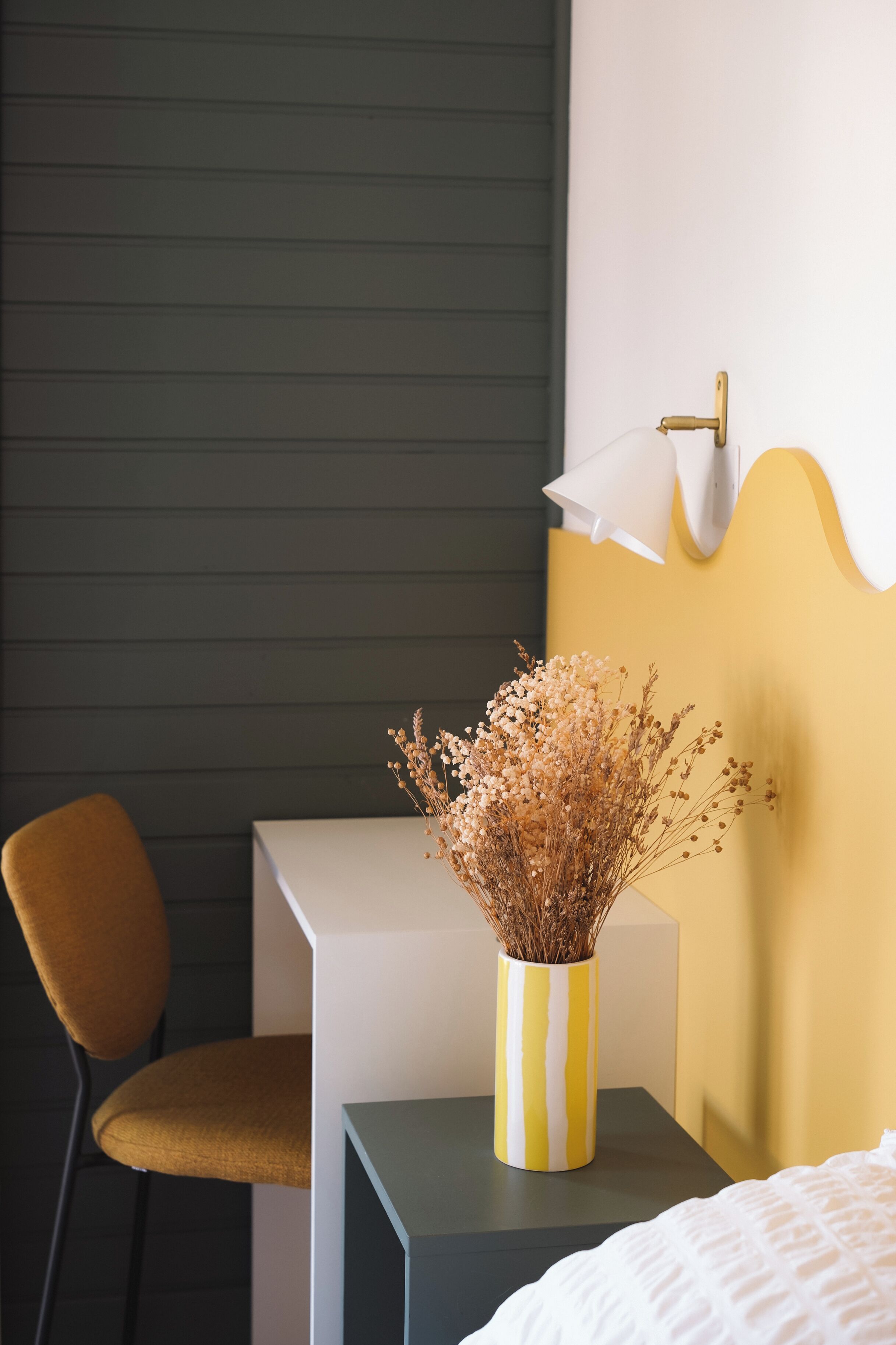 Une pièce comprenant un lit, un bureau et une chaise. Un vase contenant des fleurs séchées est posé sur le bureau.