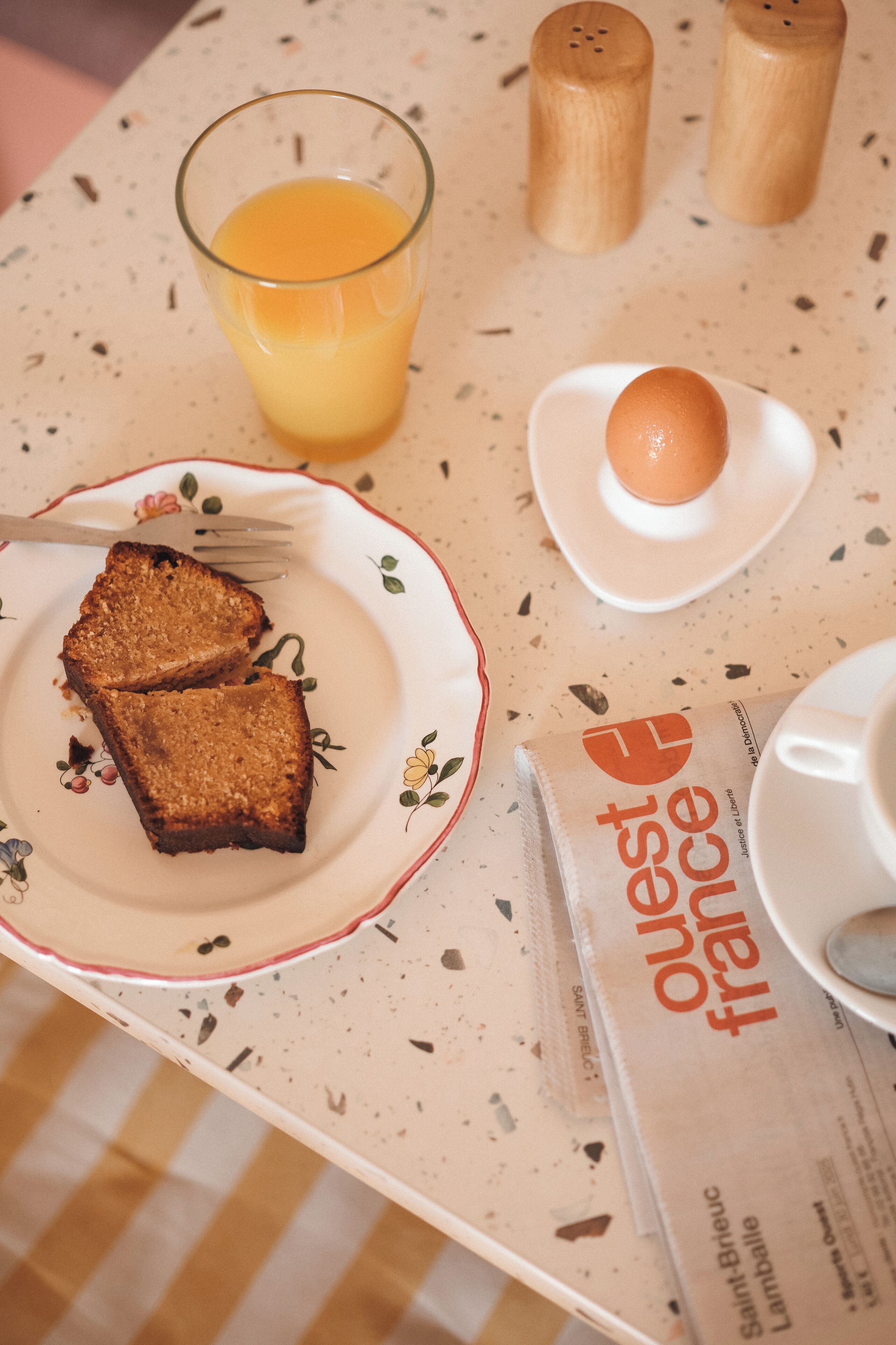 Una mesa de desayuno con tostadas, un vaso de zumo de naranja, un plato con un huevo, saleros y pimenteros, y un periódico.
