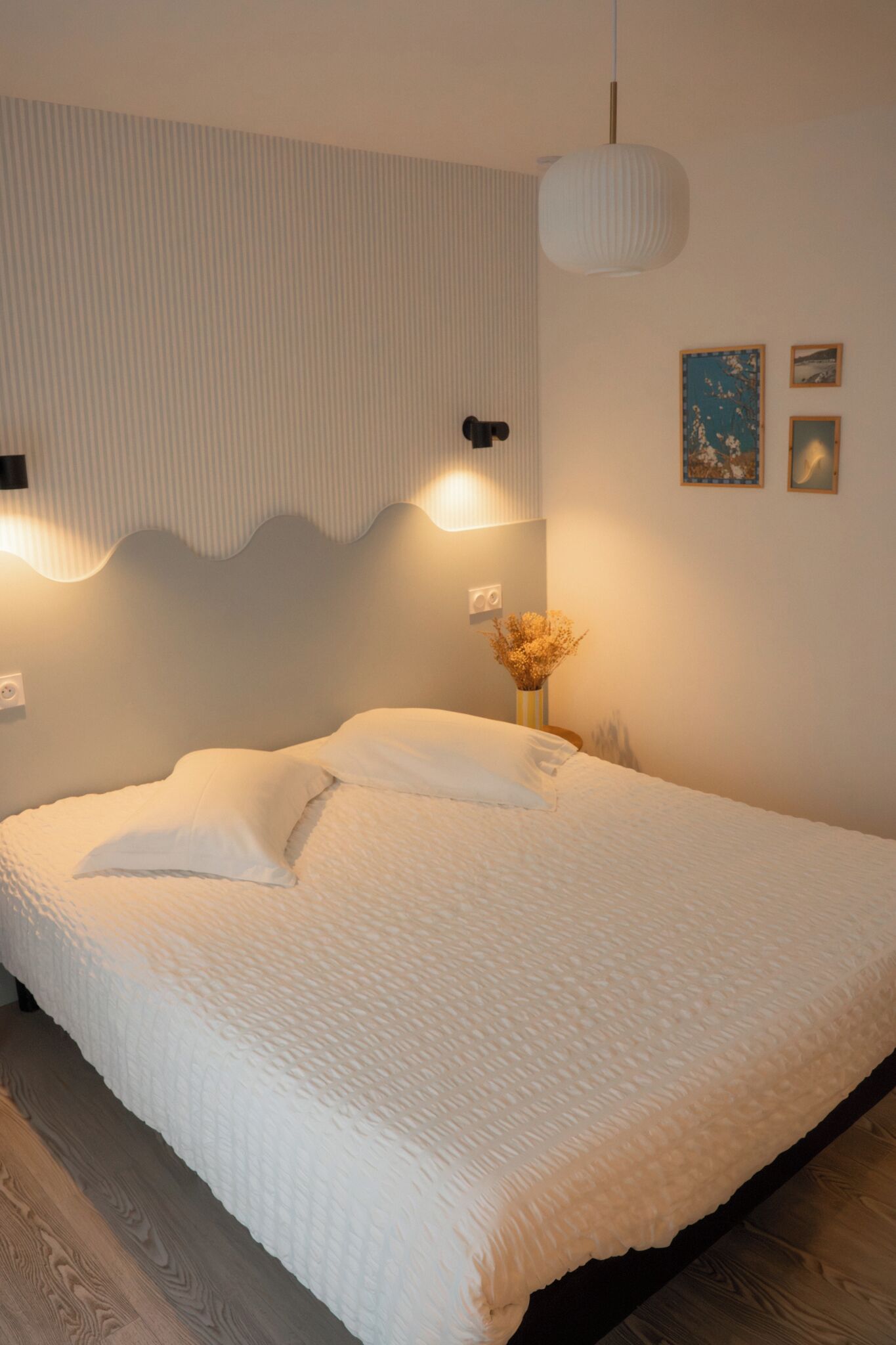 Un llit ben fet amb un edredó blanc texturat, una làmpada de paret i un petit vas amb flors seques damunt una taula de nit.