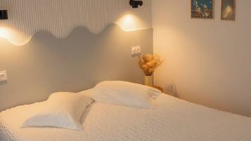 A neatly made bed with a textured white duvet, a wall-mounted light fixture, and a small vase with dried flowers on a bedside table.
