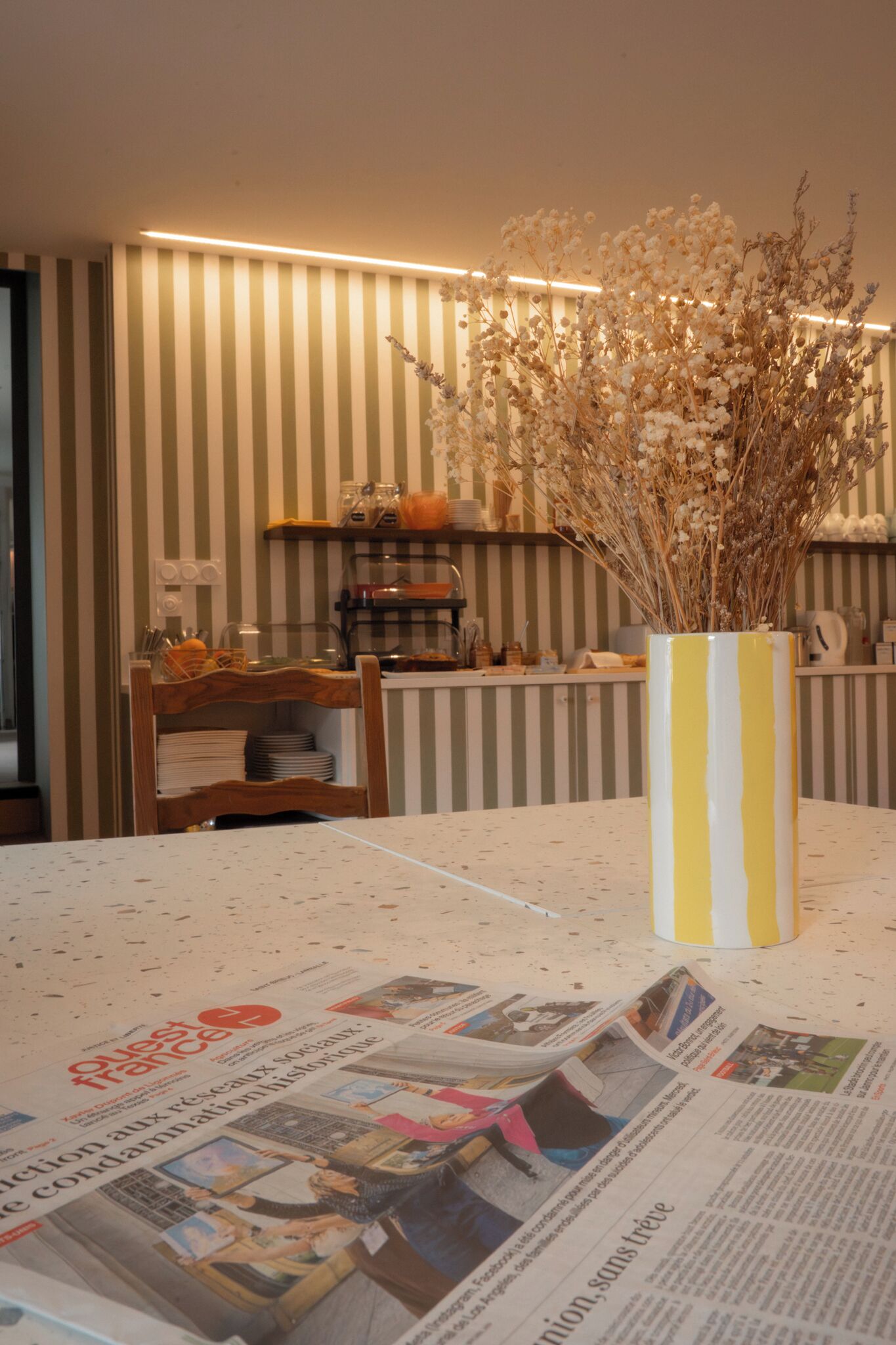 Un comedor con una pared a rayas, un jarrón con flores secas y periódicos sobre la mesa.