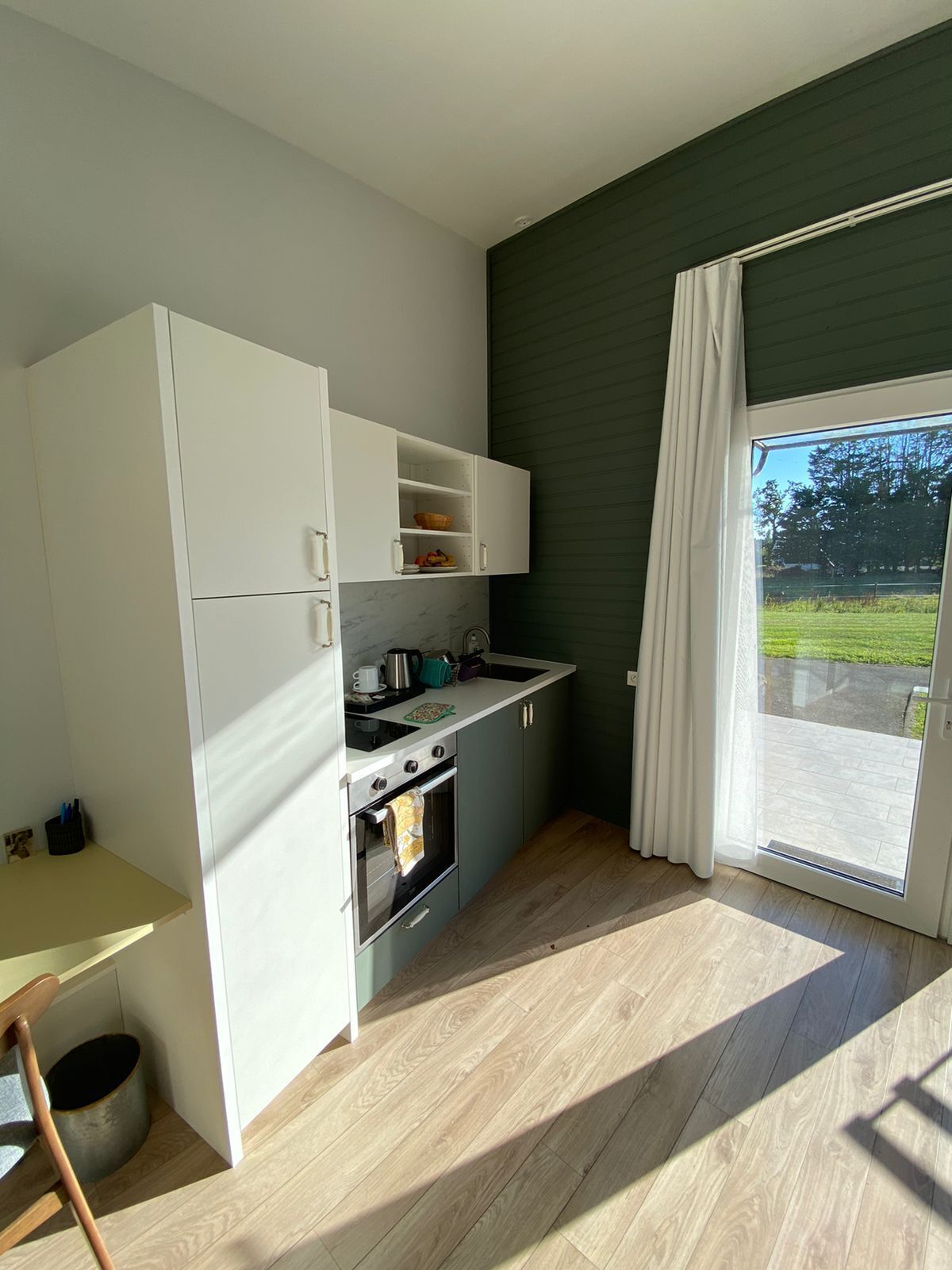 Et kompakt køkken med hvide skabe, komfur og vask. Der er et lille bord og en stol til venstre. Rummet har en stor skydedør med glas, der fører til et udendørs område.