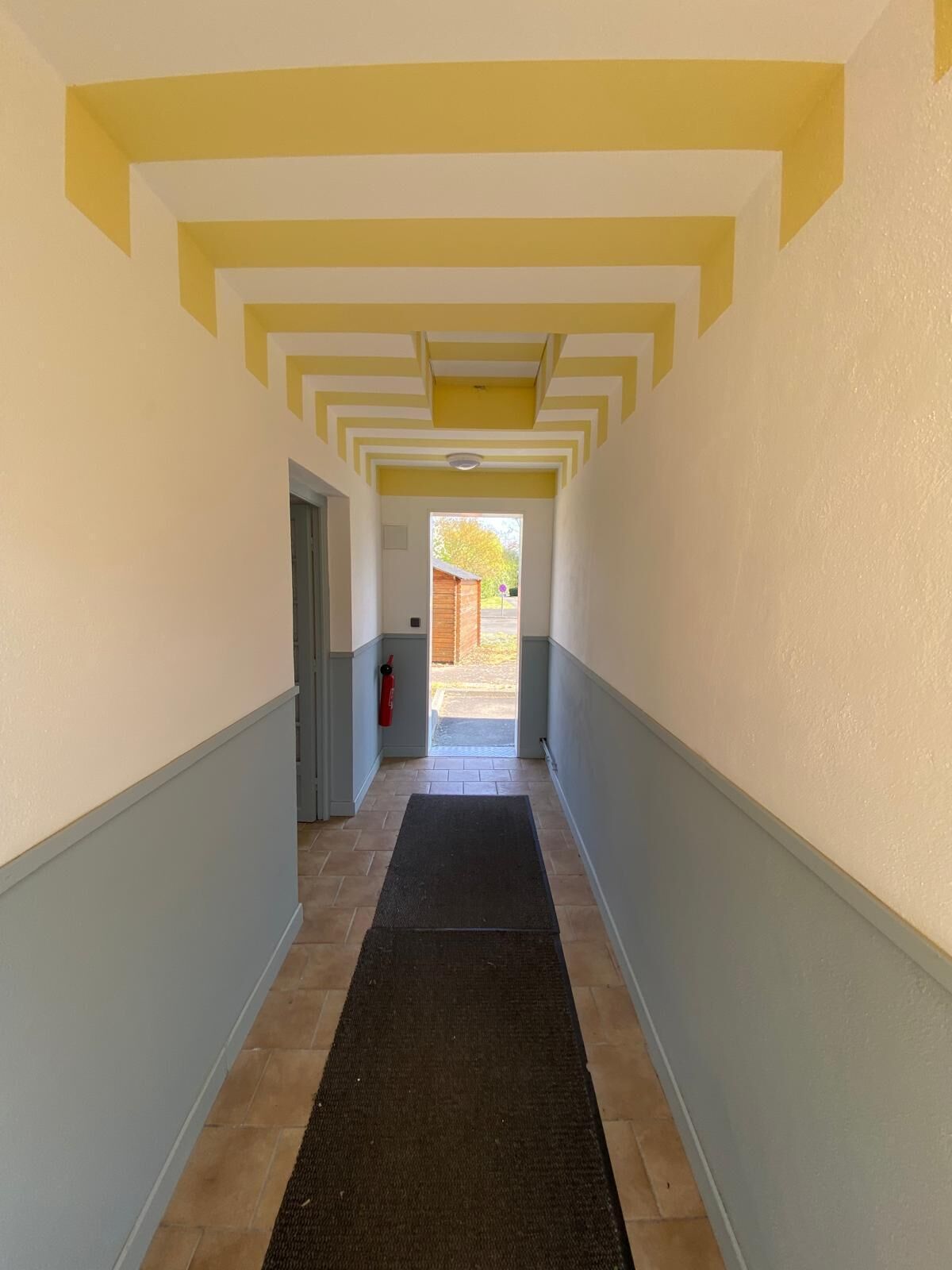 Un couloir avec un tapis noir, des murs peints en jaune et en blanc, et une porte visible au fond.