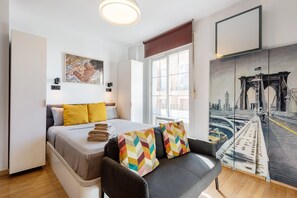 A modern hotel room with a bed, a sofa, a painting of a bridge, and a window with a view.