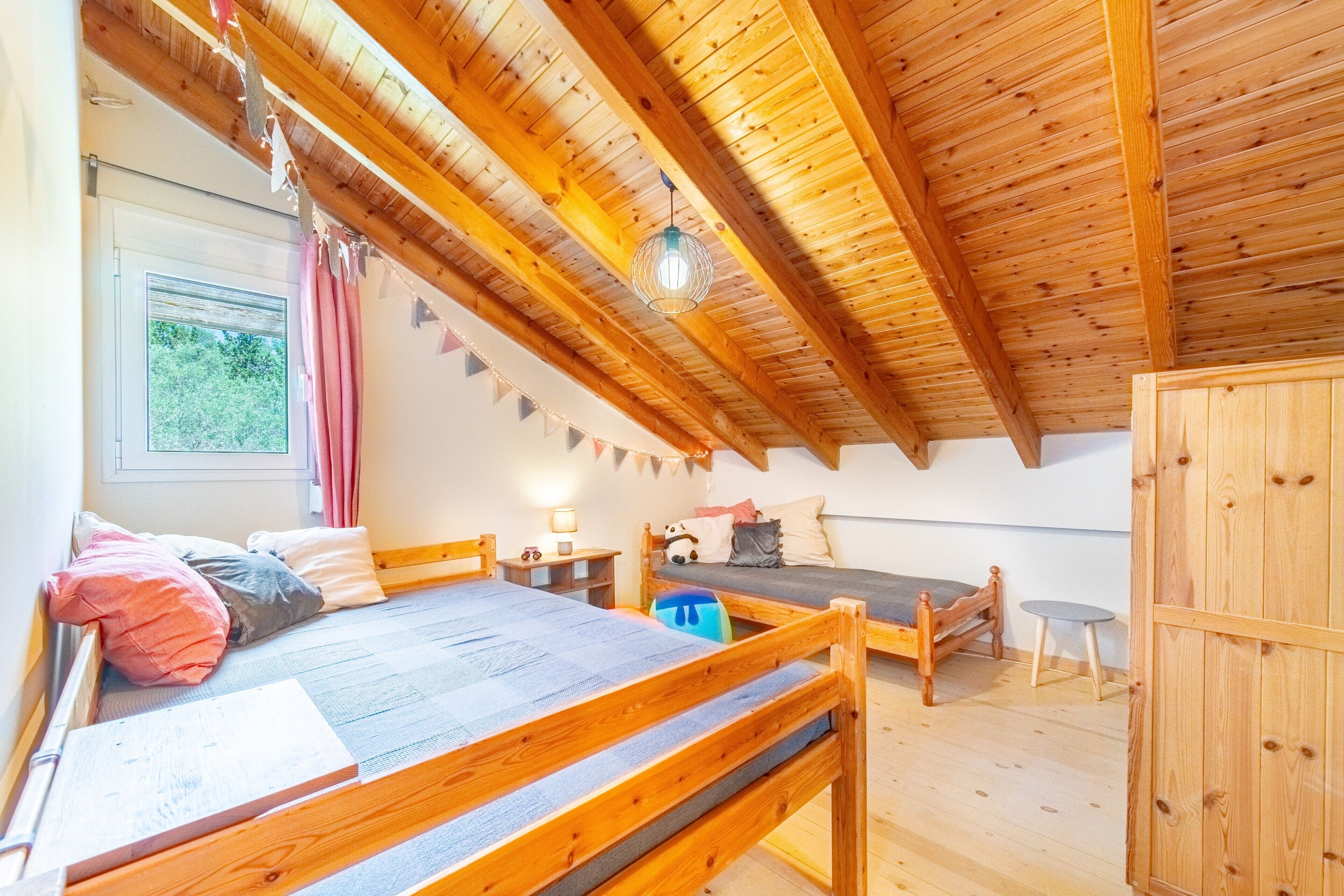 Une chambre mansardée chaleureuse avec un plafond en pente, des poutres en bois, un lit superposé, une petite table et une fenêtre avec des rideaux.