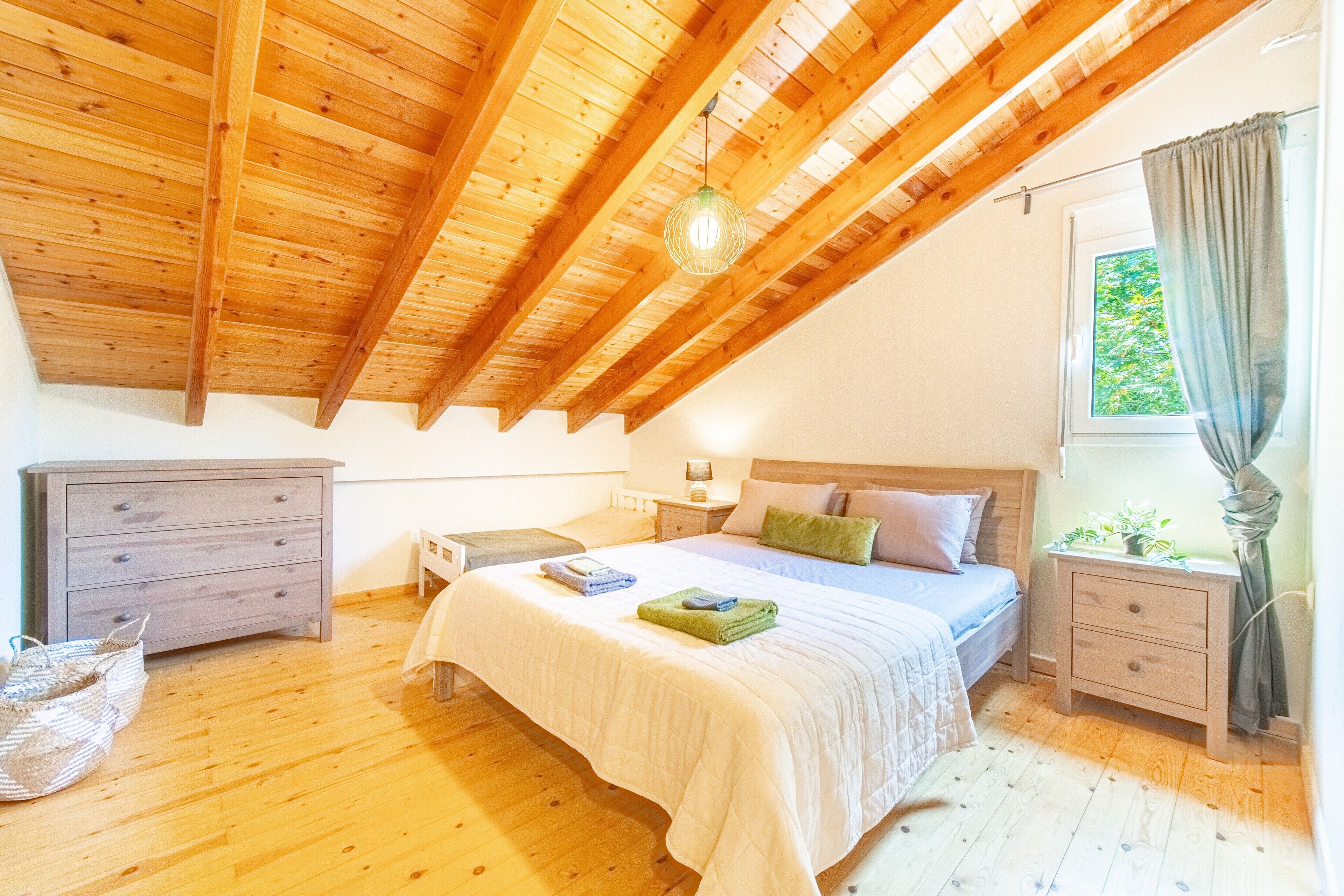 Une chambre avec un plafond en pente, un lit avec des oreillers, une table de chevet, une commode, une lampe et une fenêtre avec des rideaux.