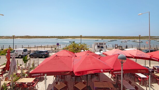 An outdoor seating area with red umbrellas, tables, and chairs, overlooking a body of water with boats.