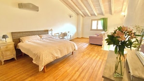 Um quarto com piso de madeira, uma cama com colcha branca, mesa de cabeceira, uma cadeira, escrivaninha e janela com cortinas.