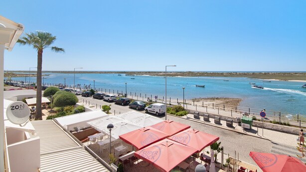 Vista costera con coches aparcados, una palmera y un restaurante frente a la playa con sombrillas rojas.