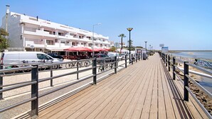 Un paseo marítimo de madera con barandilla, un edificio blanco con balcones y sombrillas rojas.