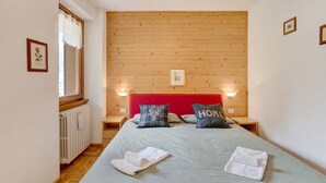 A room with a bed, two bedside tables, a window with curtains, a radiator, and a wooden headboard. - Apartment 'Chalet Holly' with Mountain View, Shared Garden and Wi-Fi (Bormio)