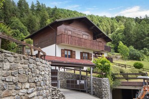 A chalet with a stone wall entrance, wooden balcony, and a view of a forested hillside.