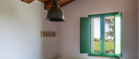 Une chambre avec un plafond en bois, un lit recouvert d’une couvre-lit rose, une fenêtre avec des volets verts et une commode blanche.