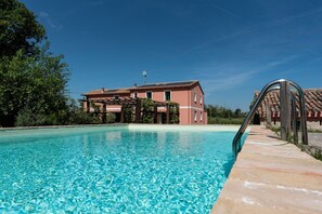 Une piscine à l’eau d’un bleu limpide, une terrasse en bois et, au fond, un bâtiment à la façade rouge.