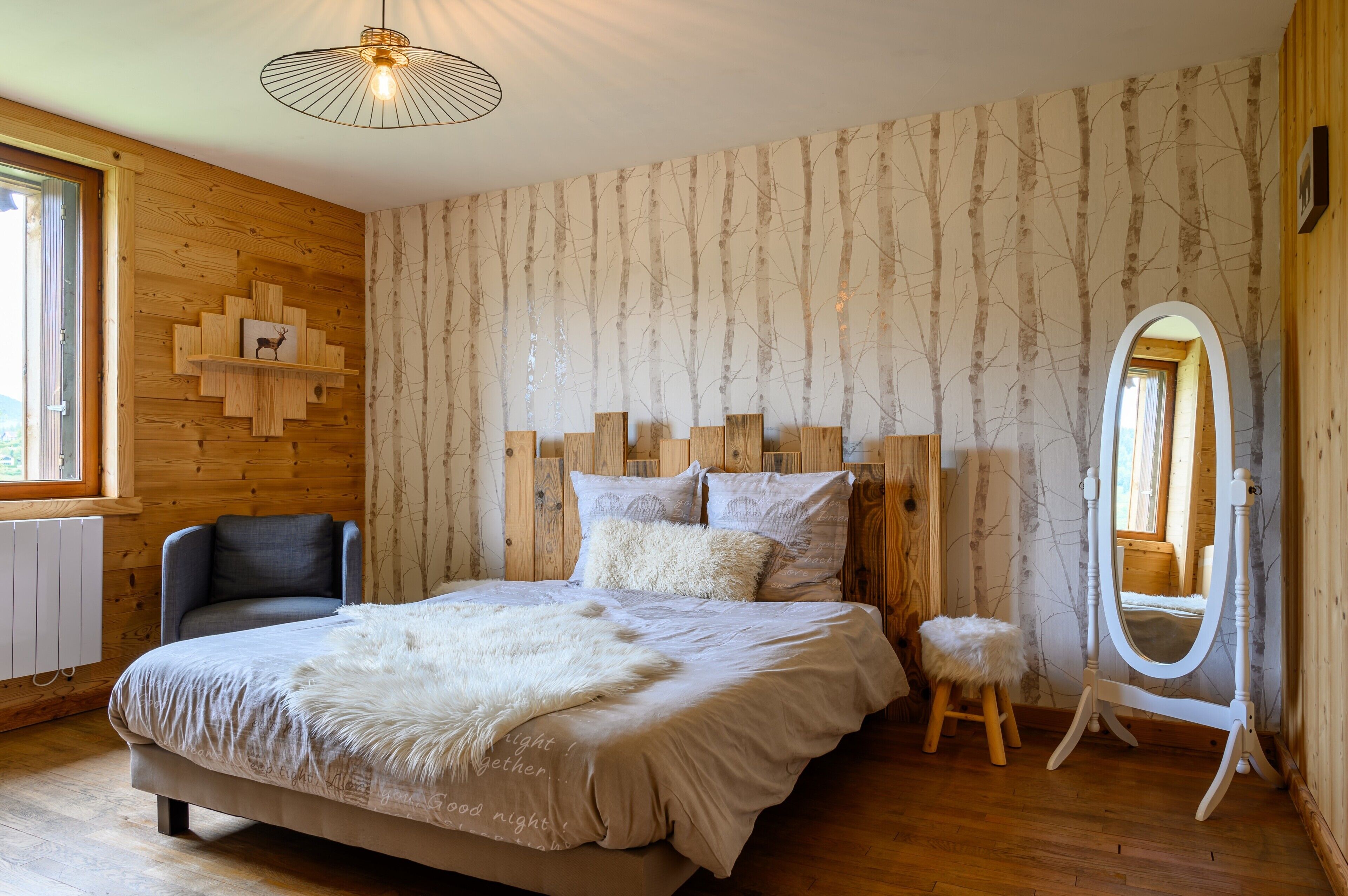 A bedroom with a wooden headboard, a bed with a white blanket, a small stool, a mirror, and a wooden shelf with a picture.