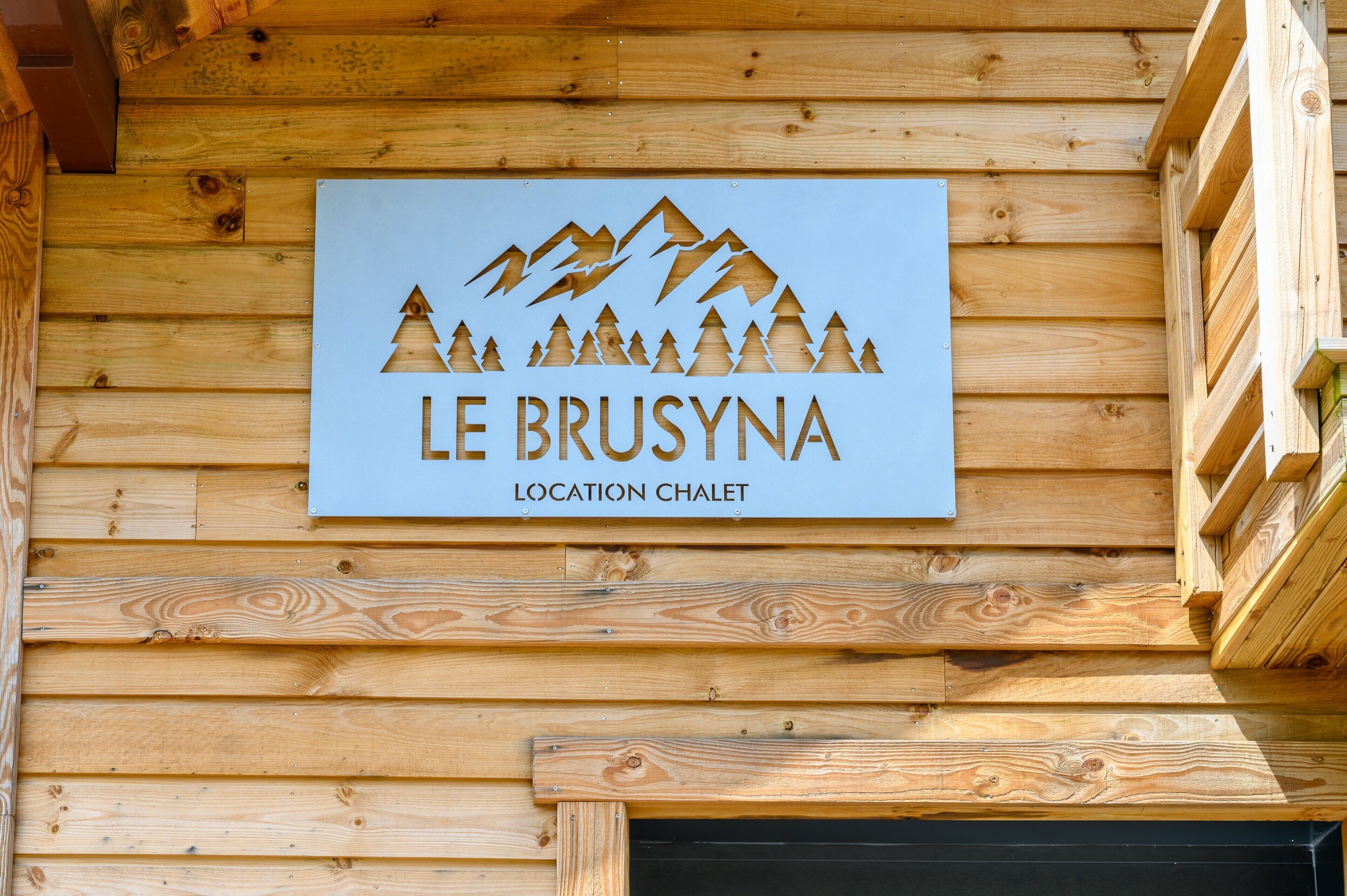 A wooden sign with a mountain and tree design, labeled 'LE BRUSYNA LOCATION CHALET.'