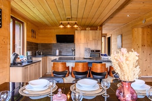 Un intérieur en bois comprenant une table à manger pour quatre personnes, une cuisine avec un grand îlot central et un téléviseur fixé au mur.