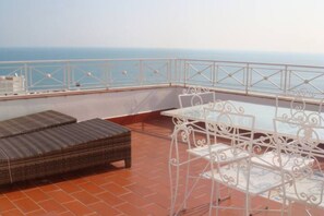 Una terraza con un sofá de mimbre, una mesa metálica blanca y sillas, con vista a un cuerpo de agua.