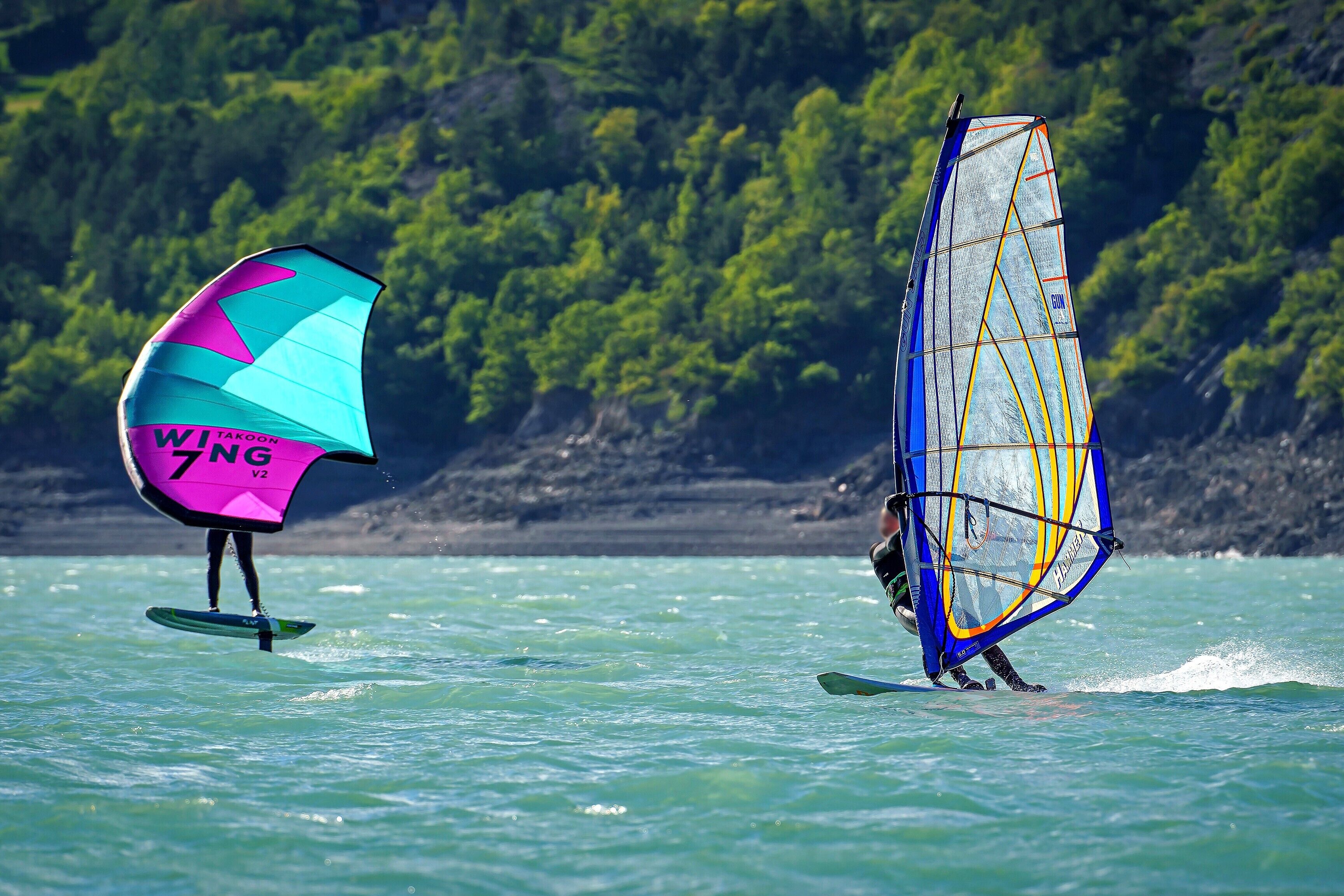 Une personne pratiquant la planche à voile avec une voile aux couleurs vives, entourée d’eau et d’une côte bordée d’une forêt.