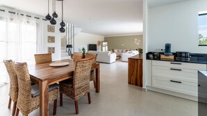 Une cuisine moderne avec des meubles blancs, une table à manger en bois et des chaises en osier, ainsi qu’un luminaire suspendu.
