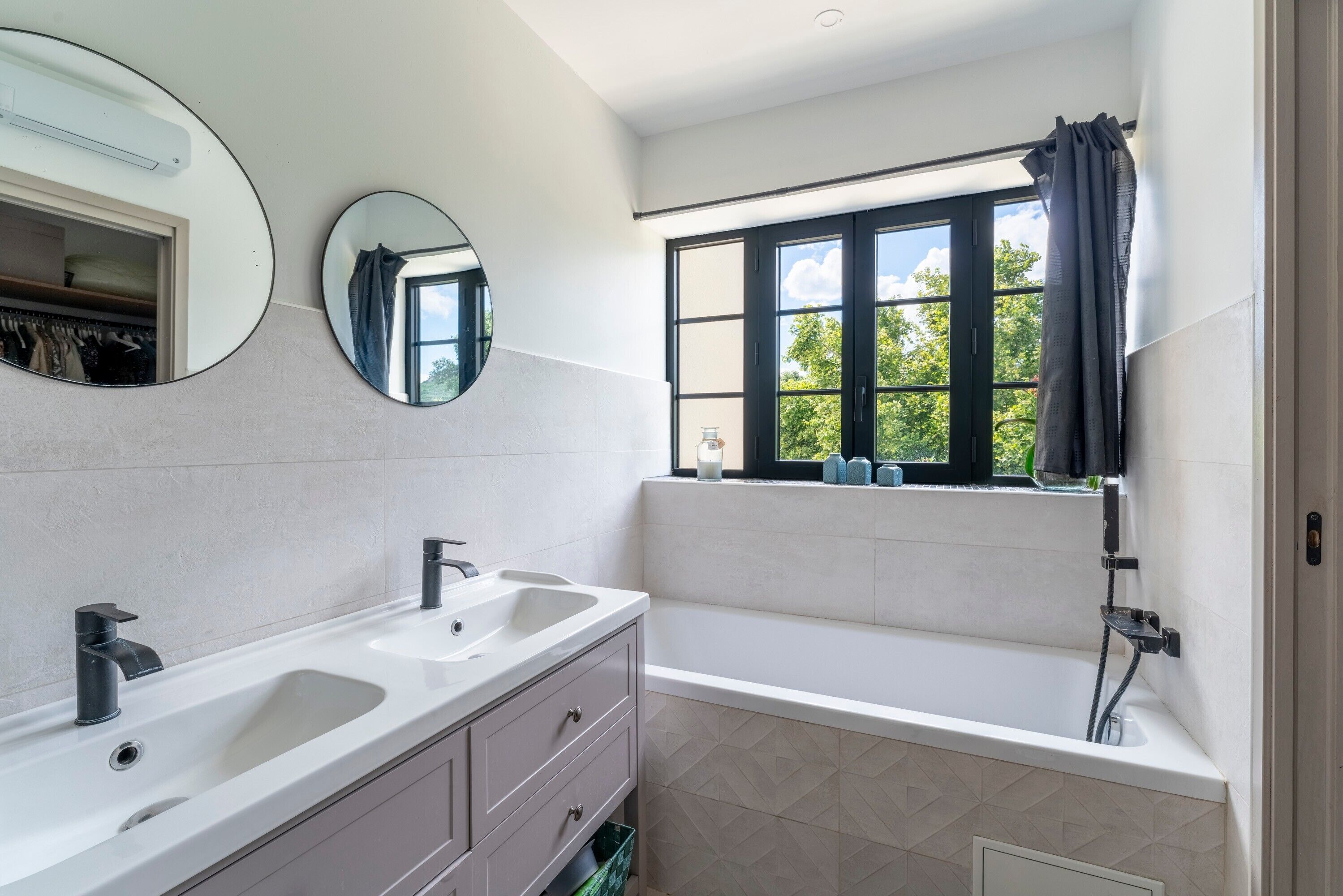A modern bathroom with a dual sink vanity, a large mirror, a bathtub, and a window with a view of greenery.