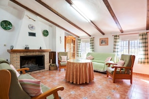 A living room with a fireplace, green sofa, wooden chairs, a round table, and a window with curtains. - Holiday Home 'Alojamiento Rural Puente Honda' with Mountain View, Pool & Wi-Fi (Benatae)