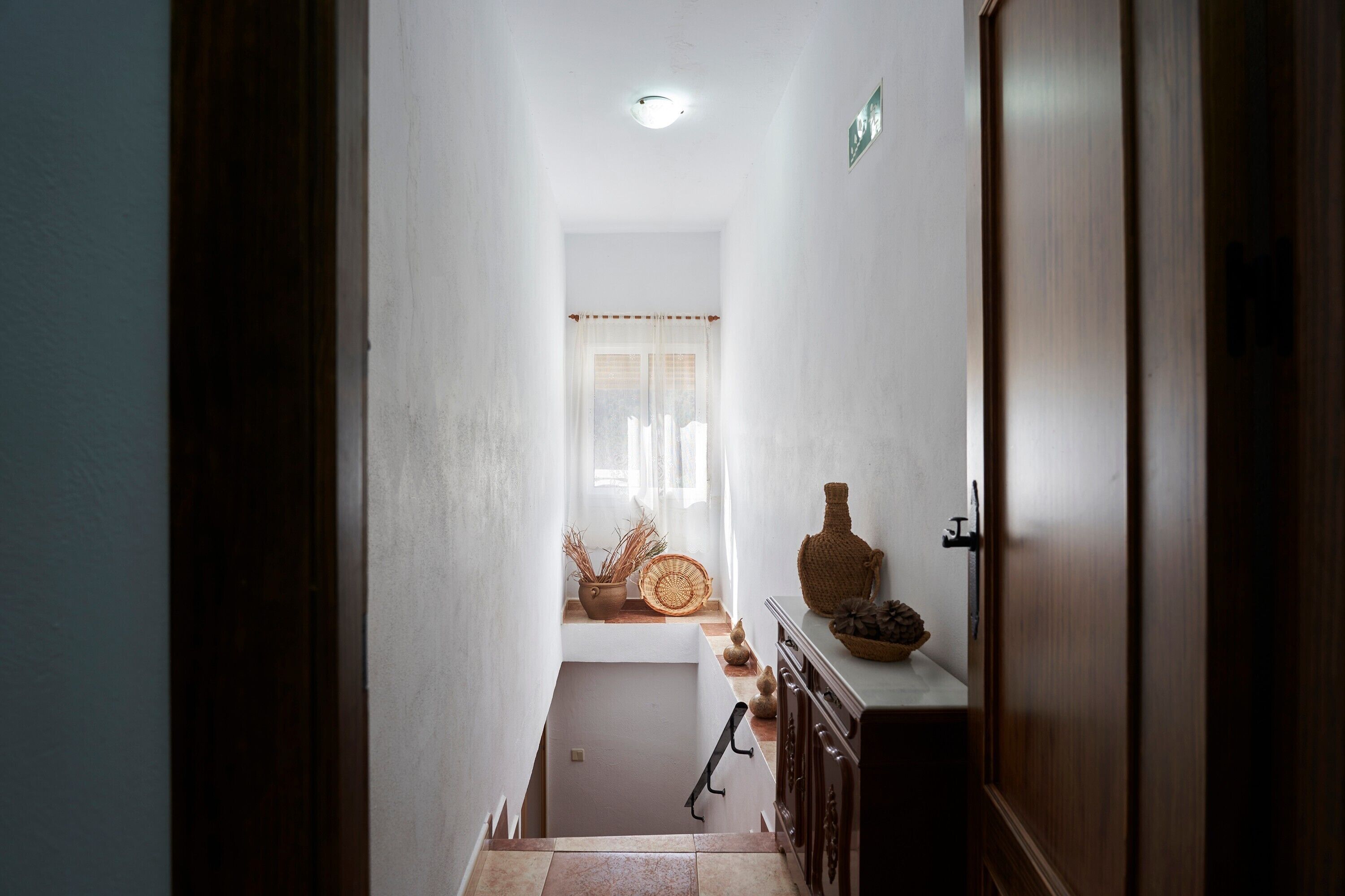 Un couloir étroit avec une porte en bois sur la droite, une petite fenêtre ornée de rideaux légers et un escalier menant à l’étage supérieur.