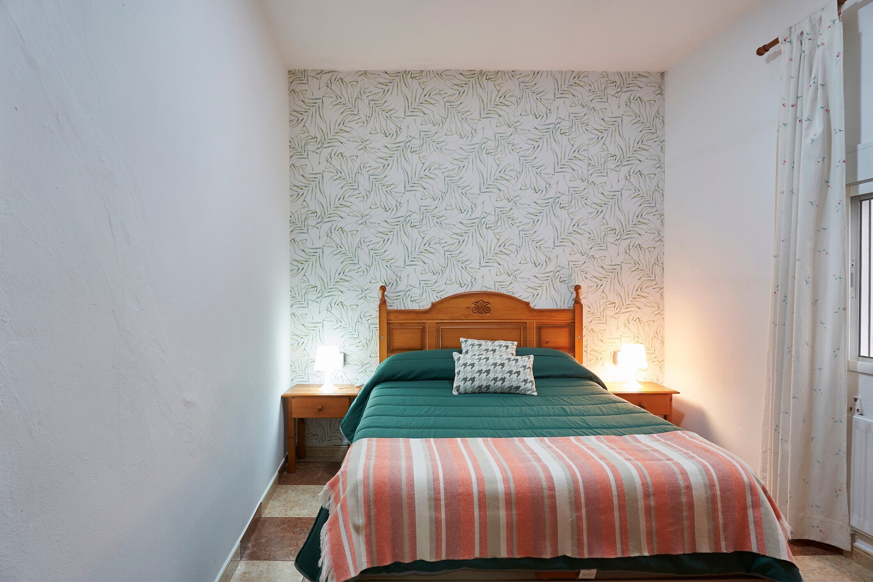 Een slaapkamer met een houten hoofdbord, een groene spreideken, een gestreept kleedje, twee bedlampjes en behang met een patroon.