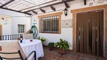Un patio con una mesa redonda, sillas, plantas en macetas, una ventana con rejas y una puerta con cortina a rayas.
