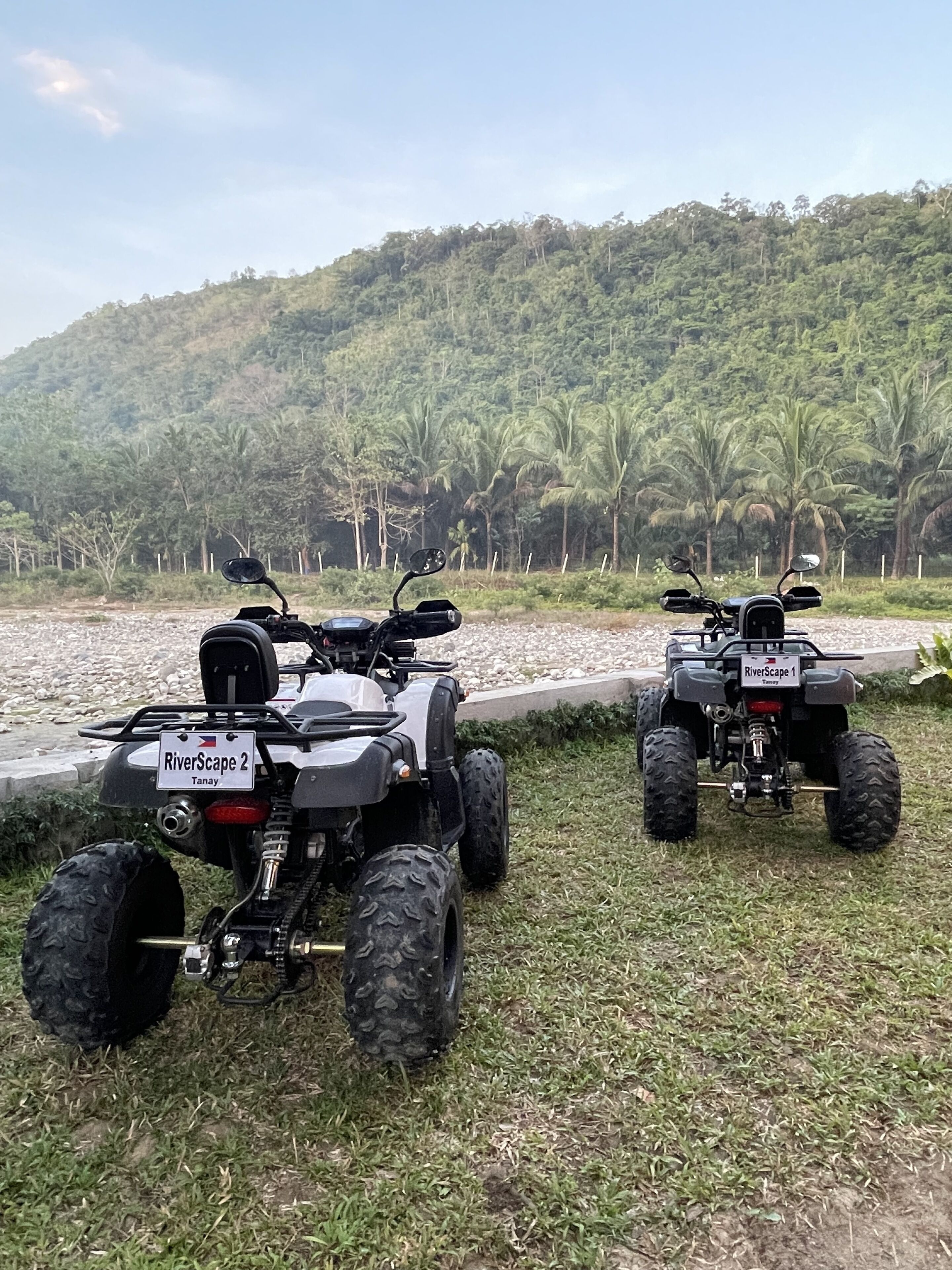 Zwei Quad-ATVs auf einer Rasenfläche in der Nähe von Gewässer, im Hintergrund ein bewaldeter Hügel.