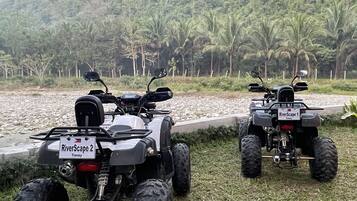 Zwei Quad-ATVs auf einer Rasenfläche in der Nähe von Gewässer, im Hintergrund ein bewaldeter Hügel.