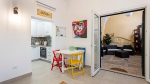 Une cuisine moderne avec des meubles blancs, une table à manger entourée de chaises rouges et jaunes, et un tableau abstrait aux couleurs vives accroché au mur.