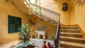 Un escalier orné d’une rampe en fer forgé, un évier et des plantes en pot dans une cour baignée de soleil.