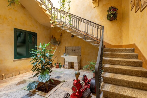 Un escalier orné d’une rampe en fer forgé, un évier et des plantes en pot dans une cour baignée de soleil.