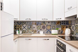 Une cuisine avec des meubles blancs, un pare-feu en carreaux, un plan de travail, un grille-pain, une bouilloire et un réfrigérateur.