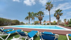 Área da piscina com espreguiçadeiras, palmeiras e céu azul límpido.