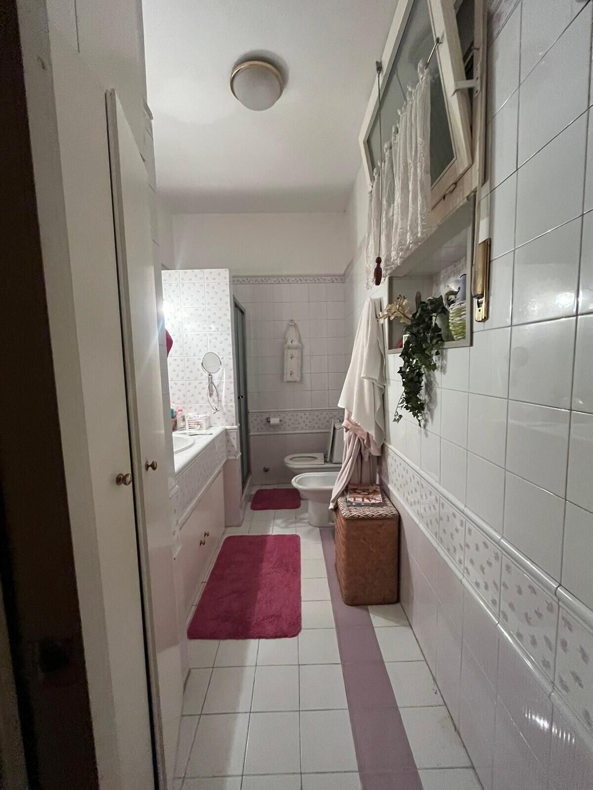 A narrow bathroom with white tiled walls, a white toilet, a sink, and a mirror. There are two pink rugs on the floor.