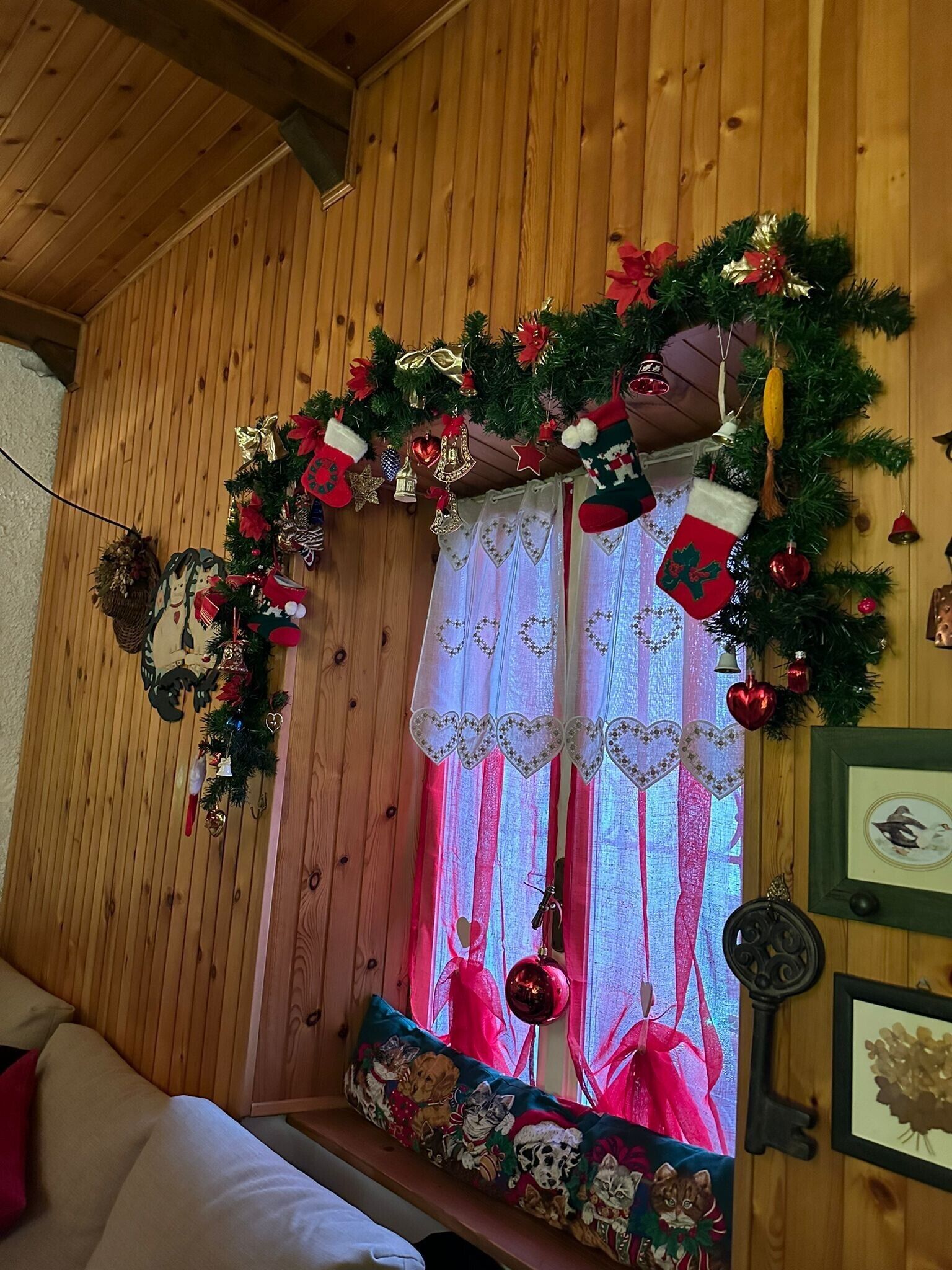 Una stanza accogliente con pareti in legno, una finestra con decorazioni festive, un cuscino con illustrazioni di gatti e un quadro appeso al muro.