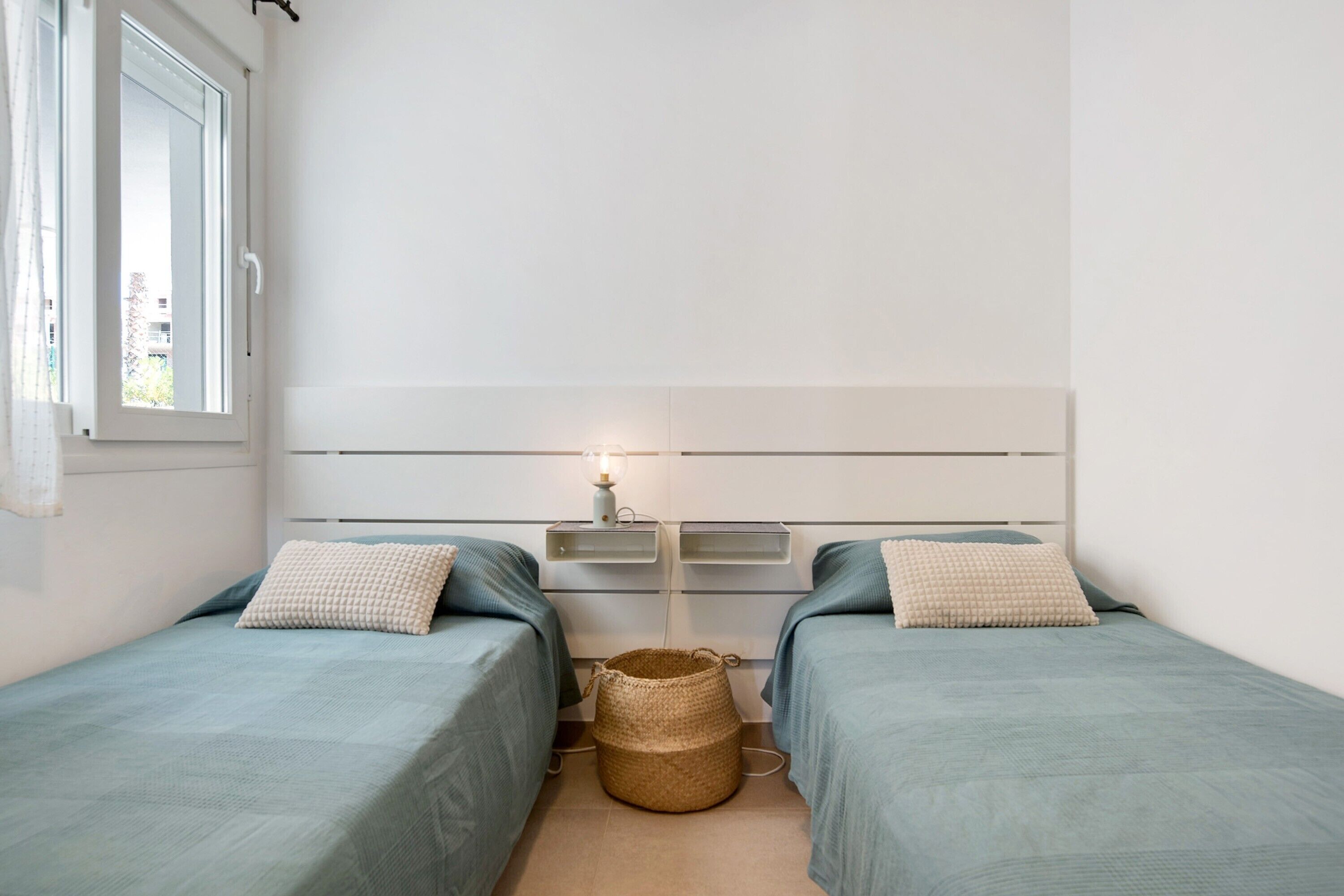 A small, clean room with two beds, a bedside table with a lamp, and a window.