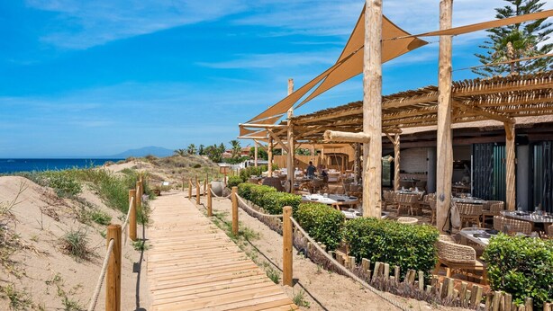 En træpromenade ved stranden, der fører til en restaurant med udendørs siddepladser og udsigt over havet.