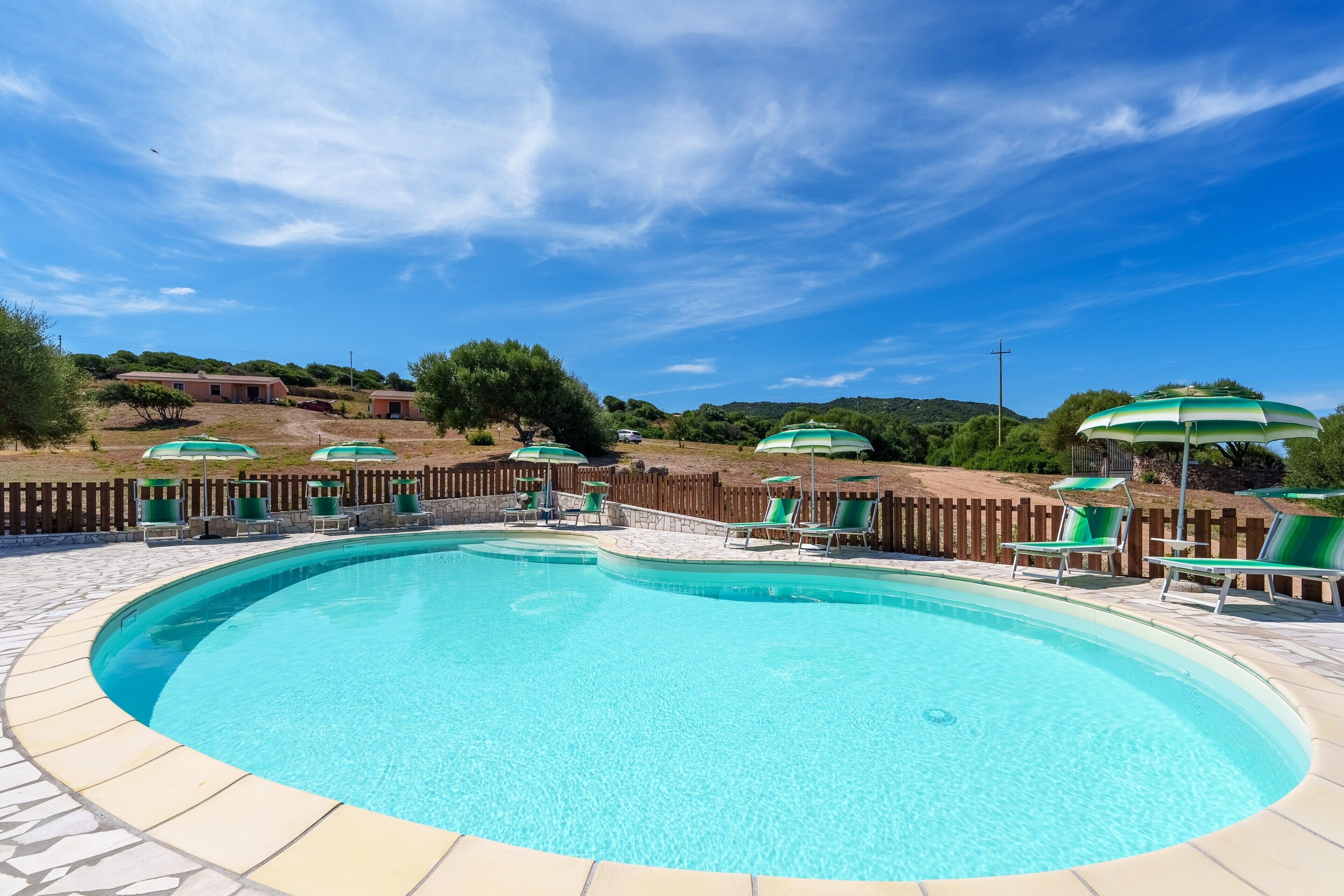 Ein Pool mit Liegestühlen und Sonnenschirmen, umgeben von einem Holenzaun und viel Grün.