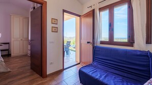 A room with a wooden floor, a blue sofa, a wardrobe, and a door leading to an outdoor area with a table and chairs. - Casa Lentischio Apartment with Sea View, Shared Pool, Terrace, and Wi-Fi (Aglientu)