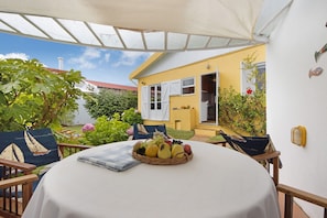Eine überdachte Terrasse mit Esstisch, im Hintergrund ein gelbes Gebäude und ein Gartenbereich mit Pflanzen und einem Stuhl.