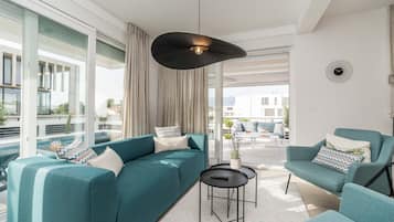 Una sala de estar moderna con sofás color esmeralda, una lámpara colgante negra y vistas de un paisaje urbano a través de amplias ventanas.