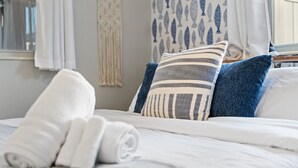 Una cama bien hecha con almohadas blancas y a rayas, una toalla blanca enrollada y una cortina azul estampada.