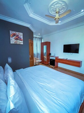 A hotel room with a large bed, a ceiling fan, a flat-screen TV, a wooden dresser, and a painting on the wall. - RK Italian Boy Hotel (Accra)